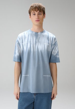 Jonge man met kort bruin haar draagt een blauwe tie-dye t-shirt en blauwe spijkerbroek, staand tegen een effen lichtgrijze achtergrond.