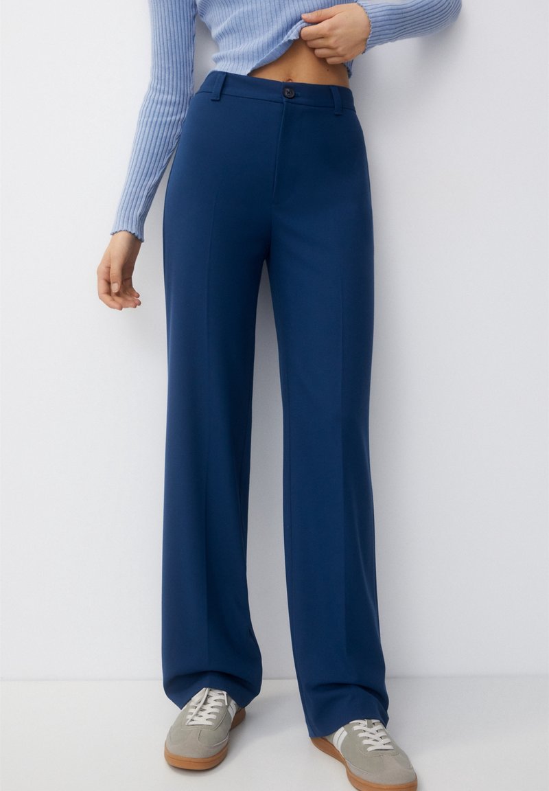 Femme portant un pantalon large bleu marine taille haute, un pull côtelé bleu clair, soulève le pull pour montrer la taille, et des baskets grises avec des lacets blancs.