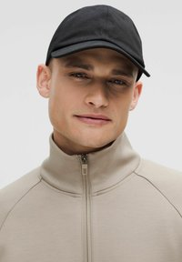 Zwarte pet met een gebogen rand, gedragen met een beige ritsjack met hoge kraag. Glad materiaal en strakke lijnen benadrukken een modern ontwerp.