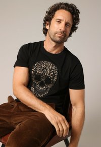 T-shirt noir en coton avec un design de crâne floral en beige. Le modèle porte un pantalon en velours côtelé marron tout en étant assis sur une chaise.