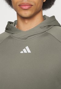 Personne portant un sweat à capuche vert olive avec le logo blanc Adidas centré sur la poitrine, montrant le bas du visage et un fond de cheveux bouclés.