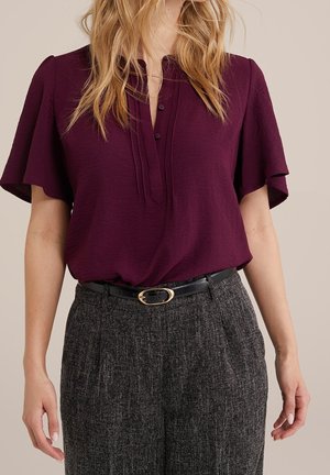 Kvinde iført en mørk blommefarvet bluse med flagrende ærmer, stoppet ned i grå teksturerede bukser med sort læderbælte og gyldent spænde.