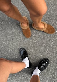 Brown suede slippers with beige faux fur trim alongside shiny black loafers with white ankle socks, set against a textured gray surface.