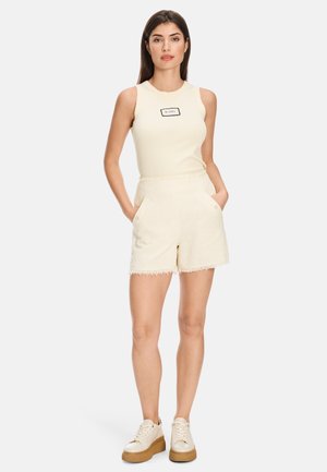 Cream tank top with “HAPPY” label and textured beige shorts. Features include side pockets and fringe hem. Paired with beige platform sneakers.