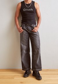 Svart ärmlös linne med texten "Crush", grå vida jeans med kontrasterande sömmar och svarta snörkängor, st ående på trägolv.