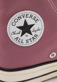 Bordové plátěné tenisky s bílou gumovou podrážkou, s kulatým logem a černou hvězdou a textem "Converse All Star". Kovové ozdobné očičky.