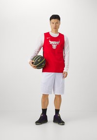 Punane korvpallivormi Chicago Bullsi logoga, valged pikad varrukad, valged šortsid ja mustad tennisetossud, hoides musta ja kollast korvpalli.
