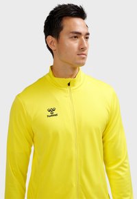 Gul atletisk lynlåstrøje med høj krave, lavet af glat stof. Har en sort logo på venstre side og et strømlinet design.