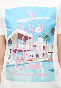 Σύγχρονη σκηνή δίπλα στην πισίνα με ξαπλώστρες, φοινικόδεντρα και ένα μινιμαλιστικό κτήριο κάτω από έναν γαλάζιο ουρανό με πουλιά και σύννεφα.