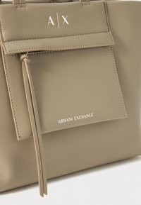 Beige szintetikus kézi táska elülső zsebbel, fehér színben nyomtatott "A/X" és "ARMANI EXCHANGE" betűkkel. Puha tapintású és struktúrált dizájn.