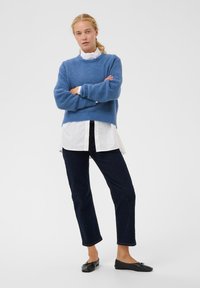 Modrý pletený svetr s texturovaným povrchem, vrstvený přes bílou košili. Nosit s tmavě modrými džínami a černými plochými botami.