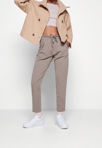 Personne portant une veste courte beige, un crop top blanc, un pantalon taupe à cordon, des chaussettes blanches et des baskets blanches, debout devant un fond uni.