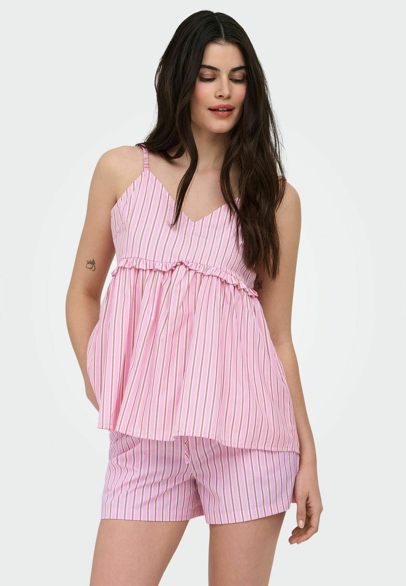 Jeune femme portant un débardeur rayé rose et blanc et un short assorti, debout, les mains dans les poches, regardant vers le bas.