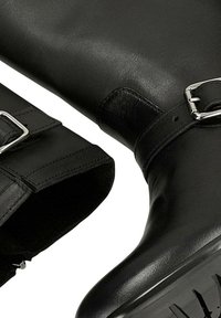Černé kožené boty se stříbrnými přezkami a robustními gumovými podrážkami, detailní záběr na oblasti kotníku a chodidla.