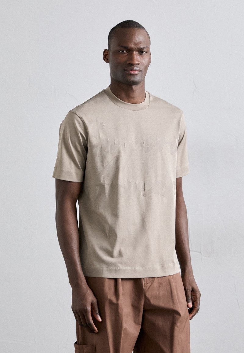 Beige kortermet t-skjorte laget av myk stoff, med et subtilt preget design på forsiden og rund hals. Brukt med brune bukser.