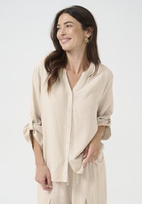 Lys beige skjorte med knapper lavet af blødt materiale, med rund halsudskæring og opkrøllede ærmer. Har en afslappet pasform og et minimalistisk design.