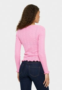 Vrouw met krullend haar, gekleed in een aansluitend roze langemouwen top met ruches aan de onderkant en donkerblauwe spijkerbroek, met haar rug naar de camera tegen een effen achtergrond.