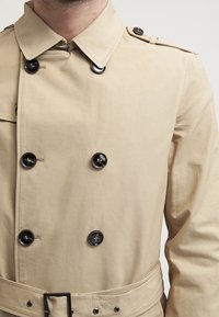 Trench coat beige con chiusura doppiopetto, bottoni neri, spalline e cintura. Tessuto liscio con un design pulito e su misura.