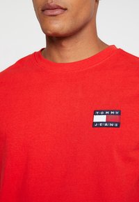 Červené bavlněné tričko s kulatým výstřihem, které má na levé straně hrudi malé vyšívané logo Tommy Jeans. Hladká textura, jednoduchý design.