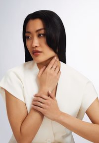 A woman in a white blouse holds her neck, wearing multiple rings—one with a small gemstone and gold bands—showcasing delicate details.