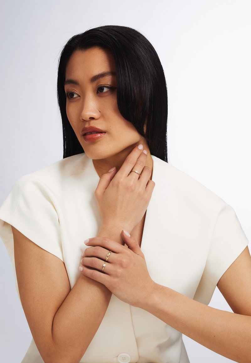 A woman in a white blouse holds her neck, wearing multiple rings—one with a small gemstone and gold bands—showcasing delicate details.