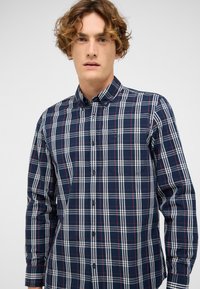 Junger Mann mit lockigem Haar, der ein kariertes Hemd in Marineblau, Weiß und Rot mit einer Brusttasche trägt, vor einem einfarbigen Hintergrund.