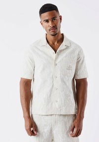 Camisa de manga corta, de color beige claro, con un patrón texturizado, bolsillo frontal y cierre de botones. Hecha de una tela ligera, diseñada para la comodidad.