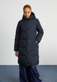 Lange zwarte pufferjack met een capuchon, voorzien van een ritssluiting en grote rechthoekige gestikte secties; gladde stof en een relaxte pasvorm.