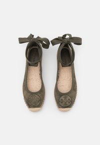 Ballerines en daim vert avec un emblème rond, bord en jute tissé et liens en ruban à la cheville. Semelle intérieure texturée avec des coutures visibles.