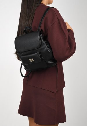 Mochila de cuero negro con un acabado texturizado, cierre de solapa plana y bolsillo frontal con acentos en hardware plateado. Lucida con un atuendo burdeos.