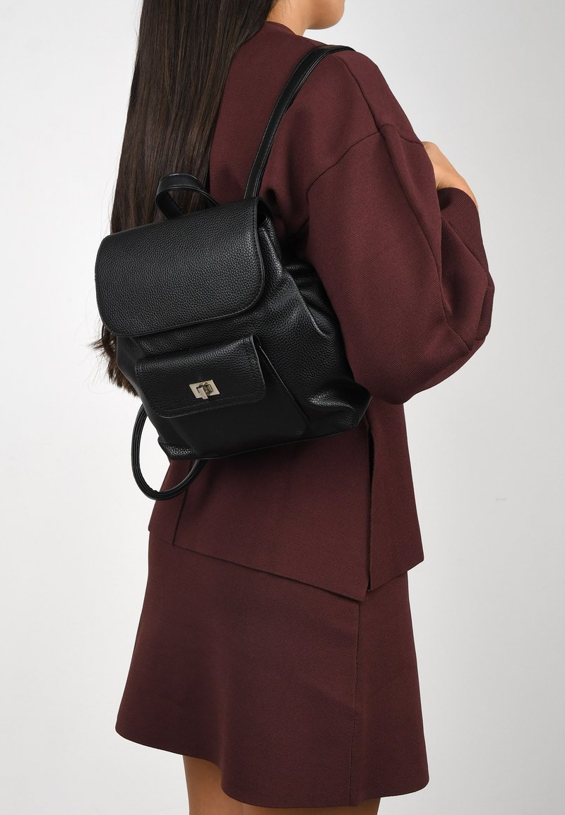 Mochila de cuero negro con un acabado texturizado, cierre de solapa plana y bolsillo frontal con acentos en hardware plateado. Lucida con un atuendo burdeos.
