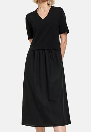 Kobieta w czarnej bluzce z krótkim rękawem i dekoltem w serek, włożonej w wysokowatą, marszczoną czarną spódnicę midi, na jednolitym tle.