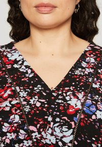 Cvetni top s črnim osnovnim barvanjem, okrašen z rdečimi, roza in modrimi vzorci. Ovratnik je globok V-izrez z poudarjenimi šivi ob šivu.