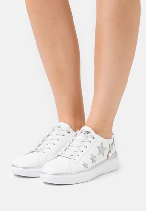 Baskets blanches à lacets avec des étoiles argentées sur les côtés, portées pieds nus sur un fond uni.