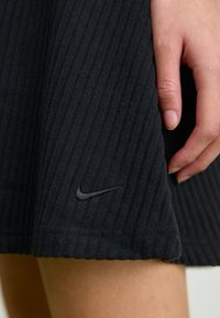 Črna rebrasta krilo iz mehkega materiala, z diskretnim Nike logotipom v spodnjem kotu. Gladka tekstura in ravna obroba.