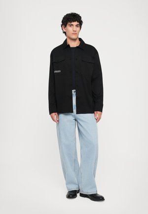 Svart knapp-up skjorte med to frontlommer, kombinert med lyseblå vide jeans. Skjorten har en subtil tekstdetalj på brystet.