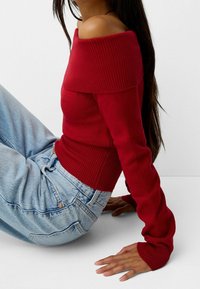 Femme assise, portant un pull rouge côtelé à épaules dénudées et un jean taille haute bleu clair, les mains posées sur le sol.