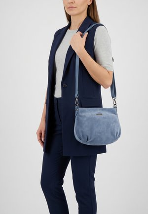 Vrouw in marineblauw vest en broek draagt een lichtblauwe suède schoudertas met verstelbare riem tegen een witte achtergrond.