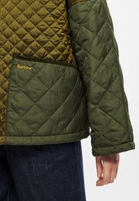 Πράσινο καπιτονέ μπουφάν με διαμάντι μοτίβο, που διαθέτει καφέ λεπτομέρεια στον ώμο και τσέπη με την ετικέτα "Barbour." Μαλακή υφή και ζεστός σχεδιασμός.