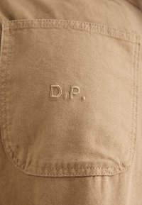 Tkanina v nijansi svetlo rjave barve z teksturirano površino, z všitom zaklopnim žepom in izvezenimi inicialkami "D.P." v usklajeni nitki.