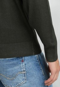 Donkergroene gebreide trui met een geribbelde zoom, gecombineerd met lichtblauwe spijkerbroek met contrasterende stiksels en een klein rood logo op de zak.