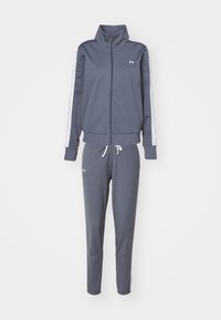 Survêtement gris foncé avec bandes blanches sur les côtés, veste zippée, pantalon à cordon et petits logos blancs Under Armour sur la poitrine et la cuisse.