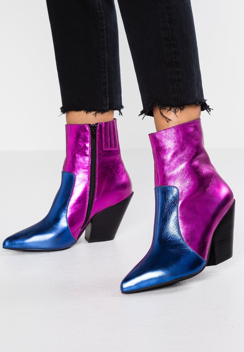 Bottines métalliques brillantes à la cheville avec bouts pointus bleus, tiges violettes, fermetures éclair latérales et talons carrés portées avec un jean noir à ourlet effiloché.