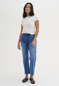 Camiseta blanca de manga corta, vaqueros azules de cintura alta con dobladillos deshilachados y zapatos negros de malla sin cordones. Diseño simple, aspecto casual.