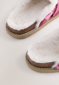 Des chaussures à enfiler roses et beiges dotées d'un col blanc duveteux, d'une semelle texturée semblable au liège et d'une tige douce semblable à de la fourrure avec des accents contrastés.