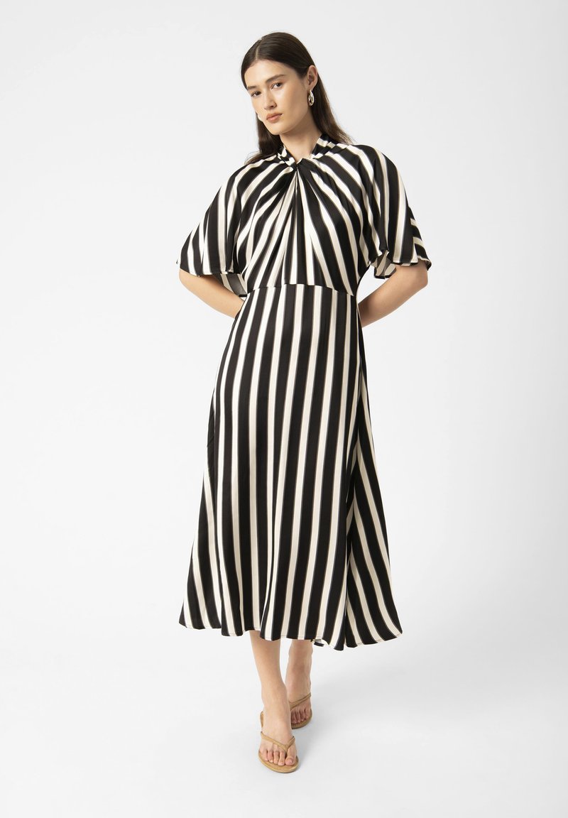 Robe à rayures noires et blanches avec des manches courtes, détail de col froncé, silhouette en A et tissu lisse. Portée avec des sandales beiges.
