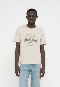 Bež pamučna majica s okruglim crnim grafičkim prikazom i tekstom "Jack & Jones" u stiliziranom fontu. Kratki rukavi i dizajn ovratnika s okruglim izrezom.
