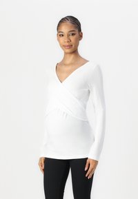 Zwangerschapstop van zachte witte stof, met een wikkeldesign, lange mouwen en een fitted vorm om een groeiende buik te accommoderen.
