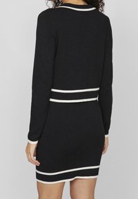 Svart klänning med långa ärmar, med vita horisontella ränder vid midjan och ärmsluten. Slät textur och figurnära design.