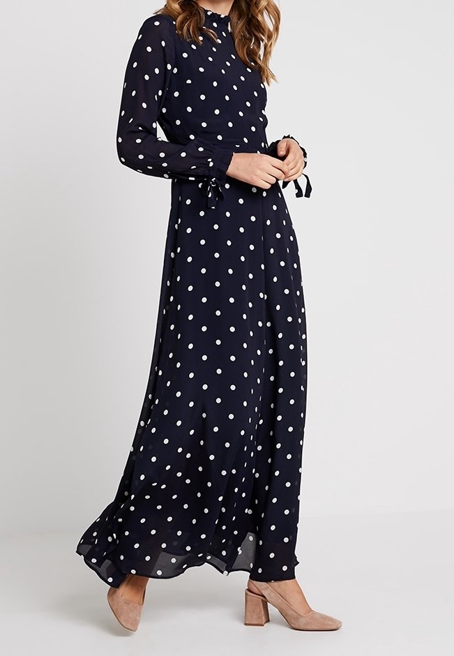 Frau trägt ein marineblaues Maxikleid mit langen Ärmeln und weißen Punkten sowie beigen Schuhe mit Blockabsatz, steht vor einem schlichten Hintergrund.